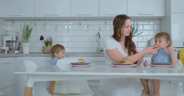 Mãe ajuda seu filho a comer um hambúrguer na cozinha — Vídeo de Stock