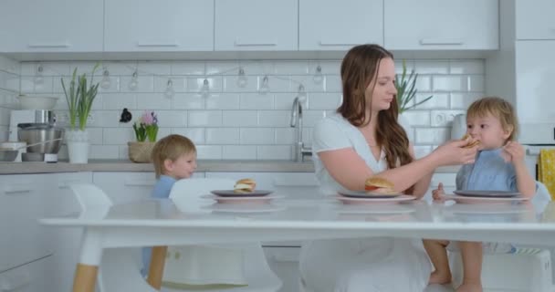 Mutter hilft ihrem kleinen Sohn, einen Burger in der Küche zu essen — Stockvideo