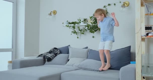 Menino bonito com cabelos brancos e camisa azul está pulando no sofá e olhando para a câmera em câmera lenta — Vídeo de Stock