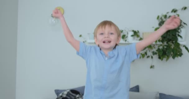 Menino bonito com cabelos brancos e camisa azul está pulando no sofá e olhando para a câmera em câmera lenta — Vídeo de Stock