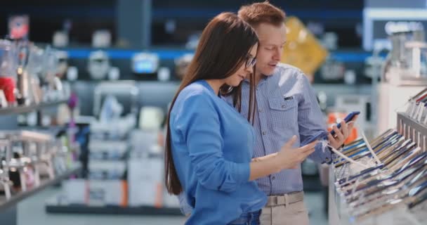 Der Mann hält sein Smartphone in der Hand und zeigt auf ein anderes Handy, das in Frauenhand ist. Innenausstattung eines Tech-Stores. — Stockvideo