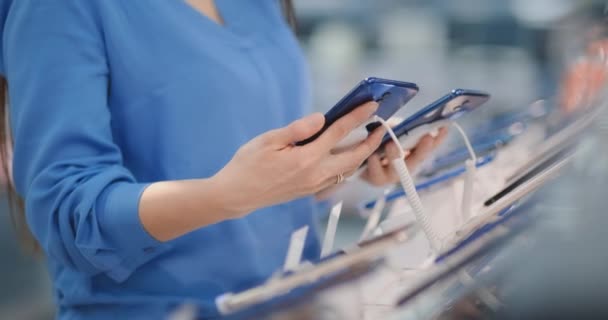 Gros plan d'une femme tenant deux nouveaux smartphones dans ses mains et choisissant le meilleur modèle de téléphone dans le magasin d'électronique moderne à acheter — Video