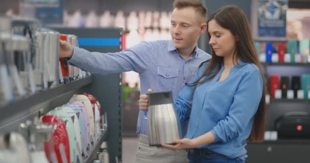 Joven pareja hombre y mujer en la tienda de electrodomésticos elige un hervidor eléctrico en el mostrador — Vídeo de stock