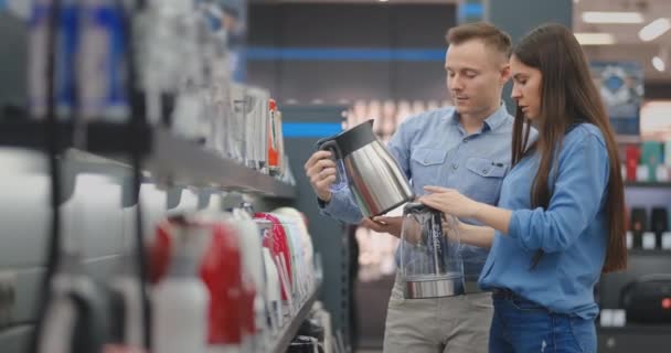 Le couple en vêtements décontractés dans le magasin d'électronique choisit une bouilloire électrique pour leur cuisine, tenant dans leurs mains et lisant et lisant les étiquettes de prix avec les caractéristiques — Video