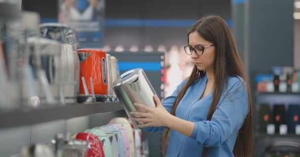 Jonge mooie brunette vrouw kiest een waterkoker te kopen. Inspecteert het apparaat, onderzoekt de prijskaartjes en de kenmerken van het model. — Stockvideo