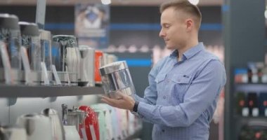 Genç ve güzel bir adam elektrikli su ısıtıcısını seçiyor. Elinde bir cihaz tutuyor, fiyat etiketlerini, modelin özelliklerini ve tasarımını inceliyor. eşyalar mağazası