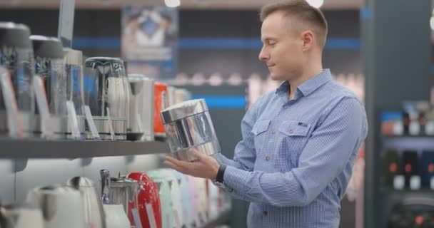 Молодой красивый мужчина выбирает электрический чайник для покупки. Держит в руках устройство, изучает ценники, характеристики и дизайн модели. магазин бытовой техники — стоковое видео