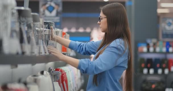 Joven hermosa morena mujer elige un hervidor eléctrico para comprar. Lleva un dispositivo en sus manos, estudia las etiquetas de precios y las características y el diseño del modelo — Vídeo de stock