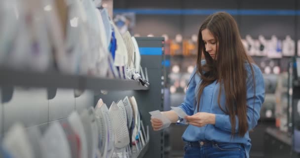 Молодая красивая женщина в рубашке с характеристиками железа изучает цены и выбирает купить утюг в магазине, стоящем у прилавка — стоковое видео