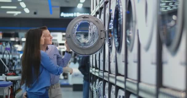 Um homem bonito e uma mulher escolhem uma máquina de lavar roupa para comprar abrir a porta e inspecionar o revestimento do tambor e a qualidade do produto. Mulher olhando dentro da máquina de lavar roupa — Vídeo de Stock