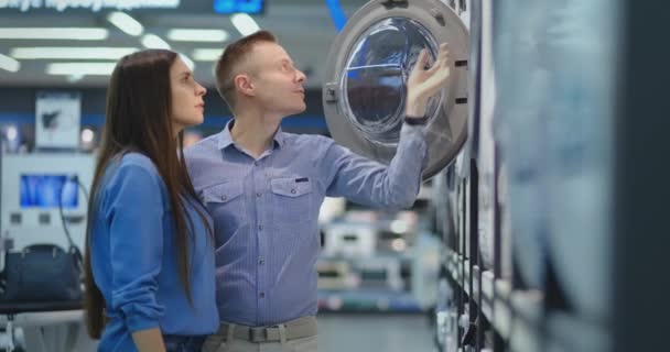 Young man and woman in appliances store choose to buy washing machine for home. Open the door looking into the drum, compare the design and characteristics of the devices. — 비디오