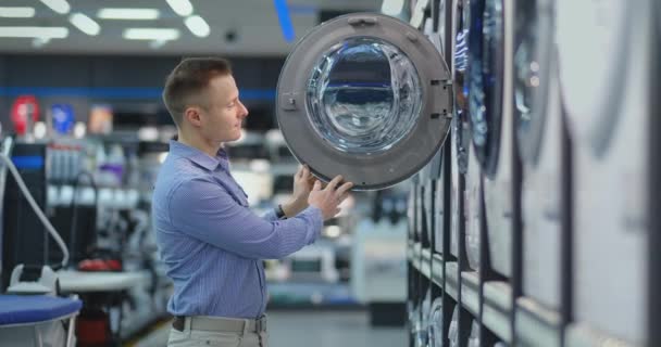 Um jovem bonito escolhe uma máquina de lavar roupa inteligente em uma loja de eletrônicos de consumo para sua nova casa. Estudos as características . — Vídeo de Stock