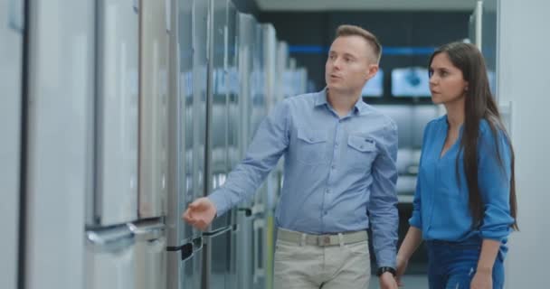 A man and a woman open the door of the new refrigerator inspect the design and quality before buying in a consumer electronics store. Read the information on the price tag and characteristics — Stock Video