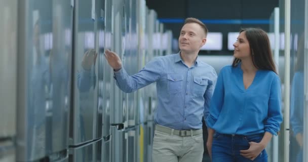Een jong stel, een man en een vrouw kiezen voor een nieuwe koelkast. Ze openen de deur en kijken naar de ruimtelijkheid en het gemak. — Stockvideo