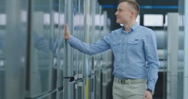 Genç yakışıklı adam satın almak için bir buzdolabı seçiyor. Kapıyı açar ve aygıtı inceler, fiyat etiketlerini ve modelin özelliklerini inceler.