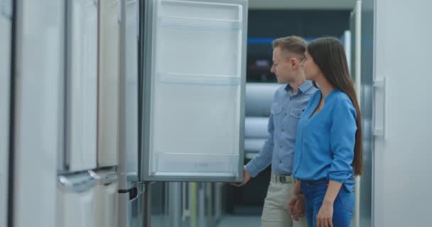 Молода подружня пара відкриває двері нового холодильника, перед покупкою в магазині побутової електроніки. Читайте інформацію про цінник і характеристики — стокове відео