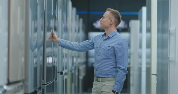 El hombre abre la puerta del refrigerador en la tienda de electrodomésticos y comparar con otros modelos para comprar la nueva casa — Vídeos de Stock