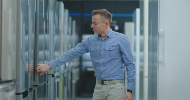 Handsome man in blue shirt open the refrigerator door in appliances store and compare with other models to buy a new home — Stock Video