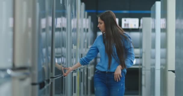 Egy fiatal nő, hogy megnyitja a hűtőszekrény ajtót tárolni készülékek és hasonlítsa össze más modellek megvenni az új házat — Stock videók