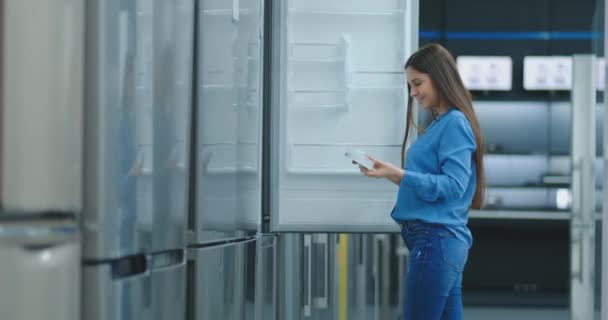 Jonge sexy brunette vrouw in shirt om de deur van de koelkast in de apparaten winkel te openen en te vergelijken met andere modellen om het nieuwe huis te kopen — Stockvideo