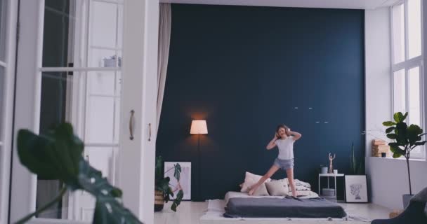 Grappige schattig klein meisje in draadloze hoofdtelefoons dansen zingen met kam en plezier hebben in vakantie ochtend springen op bed thuis in lichte slaapkamer — Stockvideo