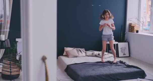 Litl lány táncolnak az ágyon, és zenét hallgat, vezeték nélküli fejhallgató. Szabadidő és modern életmód koncepció. Lány pizsama jumping ágyon reggel és mosolygós. — Stock videók