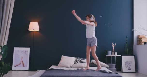 Забавная милая маленькая девочка в беспроводных наушниках танцует с расческой и весело провести время в отпуске утром прыгая на кровати дома в светлой спальне — стоковое видео