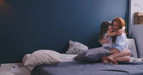 Joven madre de pelo rojo sentada en la cama abrazando a su hija sentada en pijama. Buenos días y mamá cariñosa — Vídeos de Stock