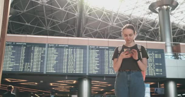 Een jonge vrouw op de luchthaven met een smartphone in de buurt van het scorebord-vertrek — Stockvideo