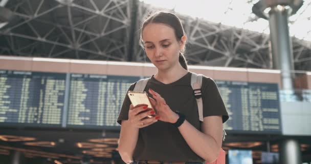 Молодая женщина в аэропорту со смартфоном возле табло-отправлений — стоковое видео