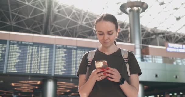 Młoda kobieta na lotnisku ze smartfonem w pobliżu tablicy wyników-wyjazdy — Wideo stockowe