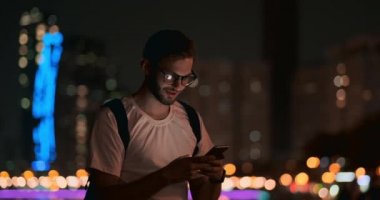 Gece şehrinin arka planına karşı gözlüklü bir adam akıllı telefon kullanarak mesaj yazıyor.
