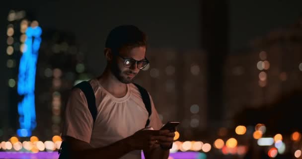 Человек в очках на фоне ночного города пишет сообщение с помощью смартфона — стоковое видео
