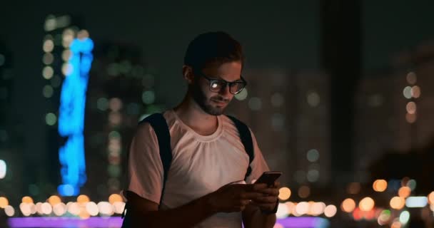 夜に眼鏡をかけた若い男がスマートフォンの画面を覗く — ストック動画