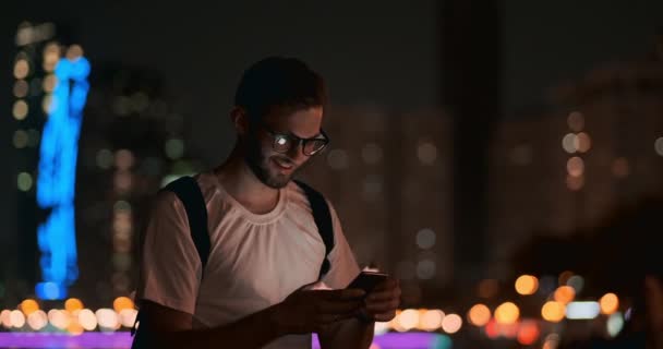 Ein junger Mann mit Brille blickt nachts in den Bildschirm eines Smartphones — Stockvideo