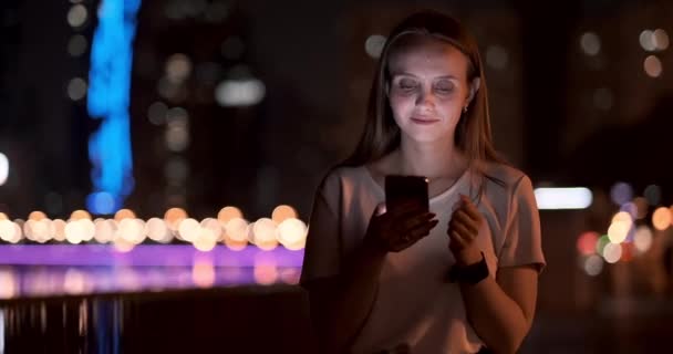 Una chica joven en el fondo de la ciudad nocturna mira en la pantalla del teléfono inteligente — Vídeos de Stock