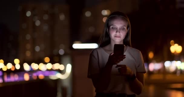 At night, a young girl holds a smartphone in her hands and looks at the screen — Stock Video