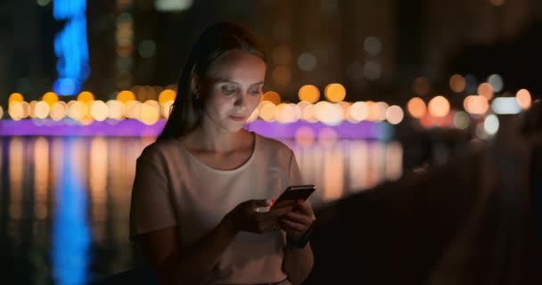 Een jong meisje op de achtergrond van de nacht stad kijkt naar het scherm van de smartphone — Stockvideo