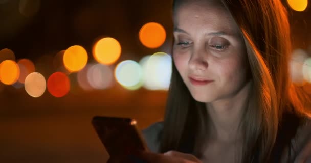 Una chica joven en el fondo de la ciudad nocturna mira en la pantalla del teléfono inteligente — Vídeos de Stock