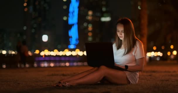 Mulher desenvolvedor senta-se no parque à noite na cidade e escreve código olhando para a tela do laptop — Vídeo de Stock