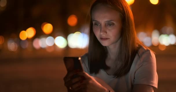 Chica joven en la ciudad nocturna escribe mensajes de texto en un teléfono inteligente — Vídeos de Stock