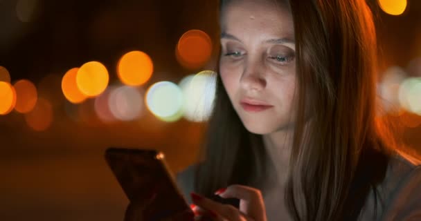 Chica joven en la ciudad nocturna escribe mensajes de texto en un teléfono inteligente — Vídeos de Stock