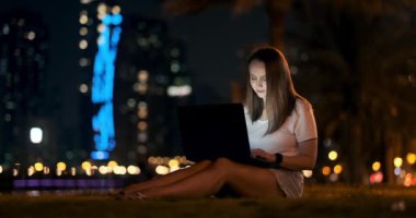 Genç bir kadın yaz gecesi çimenlerde dizüstü bilgisayarıyla oturuyor.