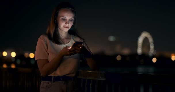 Una giovane ragazza sullo sfondo della città notturna guarda nello schermo dello smartphone — Video Stock