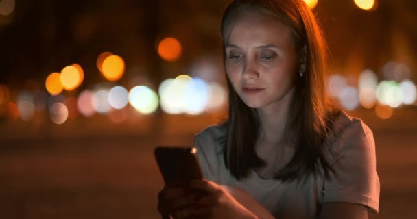 Por la noche, una joven sostiene un teléfono inteligente en sus manos y mira a la pantalla — Vídeos de Stock