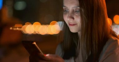 Gece şehrinin arka planında genç bir kız akıllı telefon ekranına bakıyor
