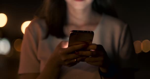 Por la noche, una joven sostiene un teléfono inteligente en sus manos y mira a la pantalla — Vídeos de Stock