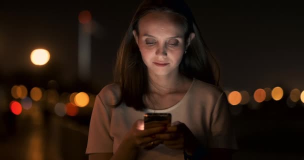 At night, a young girl holds a smartphone in her hands and looks at the screen — Stock Video