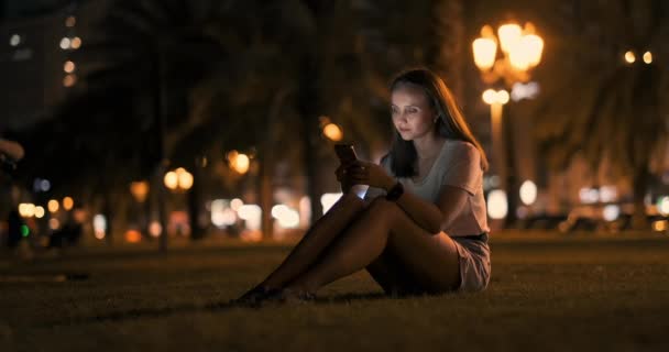 Una chica joven en el fondo de la ciudad nocturna mira en la pantalla del teléfono inteligente — Vídeos de Stock