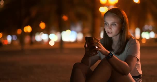 Uma jovem no fundo da cidade noturna olha para a tela do smartphone — Vídeo de Stock